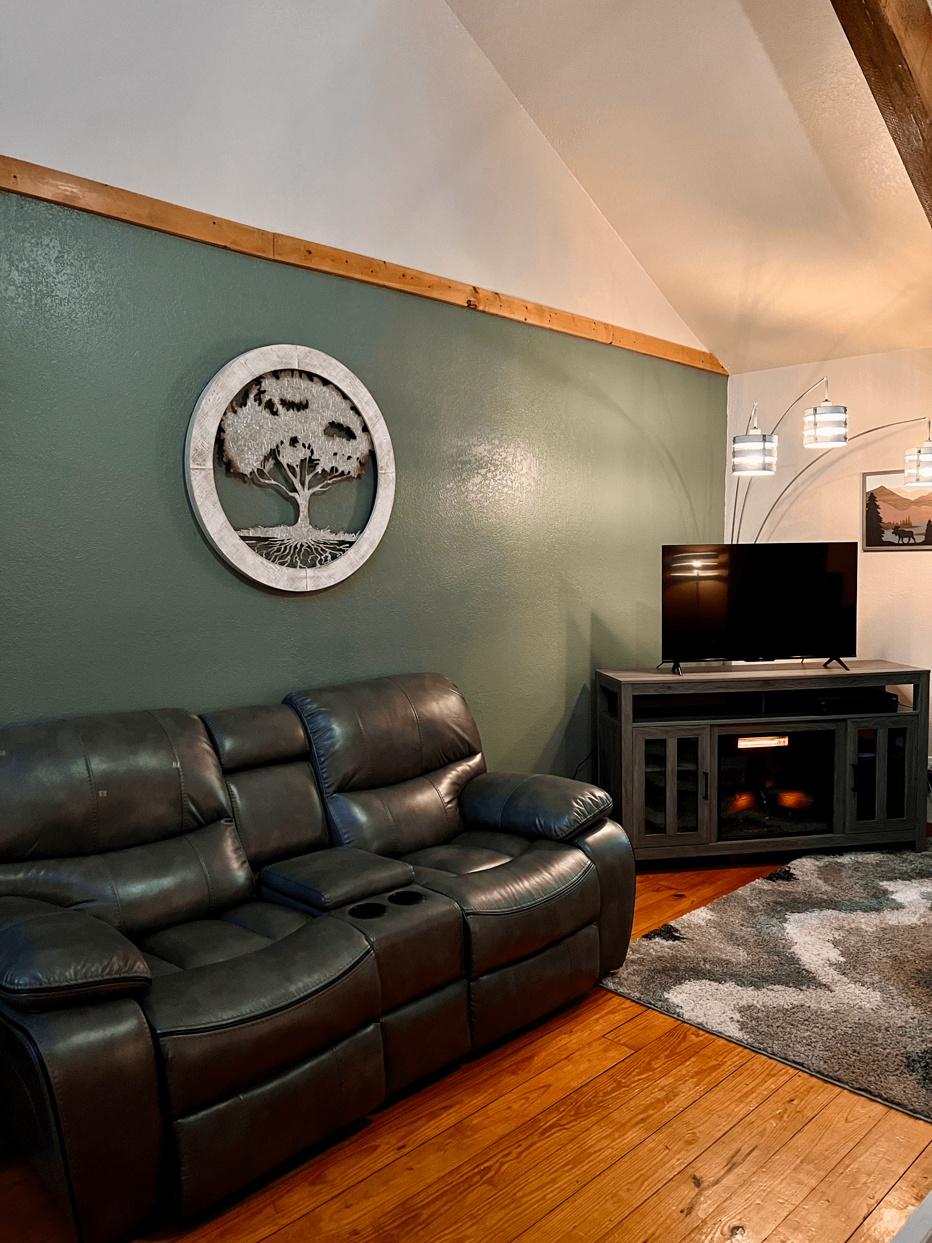 Dark leather sofa, hardwood floors with plush area rug, TV console with flat-screen TV, and tree sculpture hanging on wall