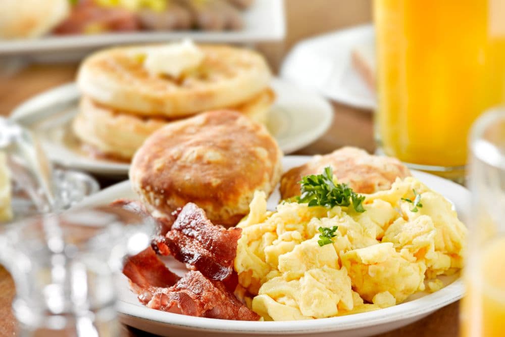 Plates of breakfast food, eggs, bacon, English muffin, waffles and orange juice