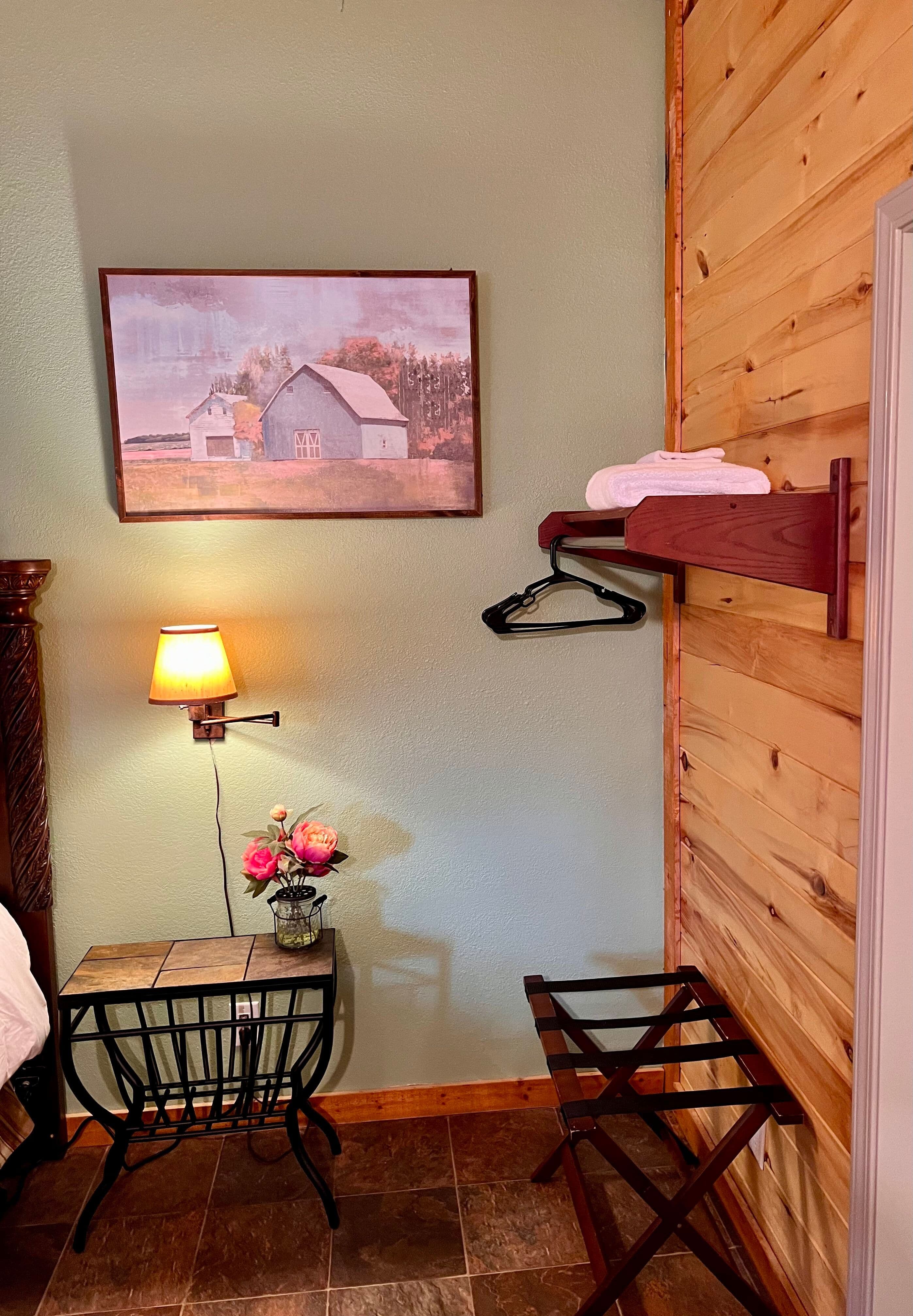 Metal nightstand with wood top and wall-mounted light and painting of country scene above, suitcase stand, and shelf with clothes hangers