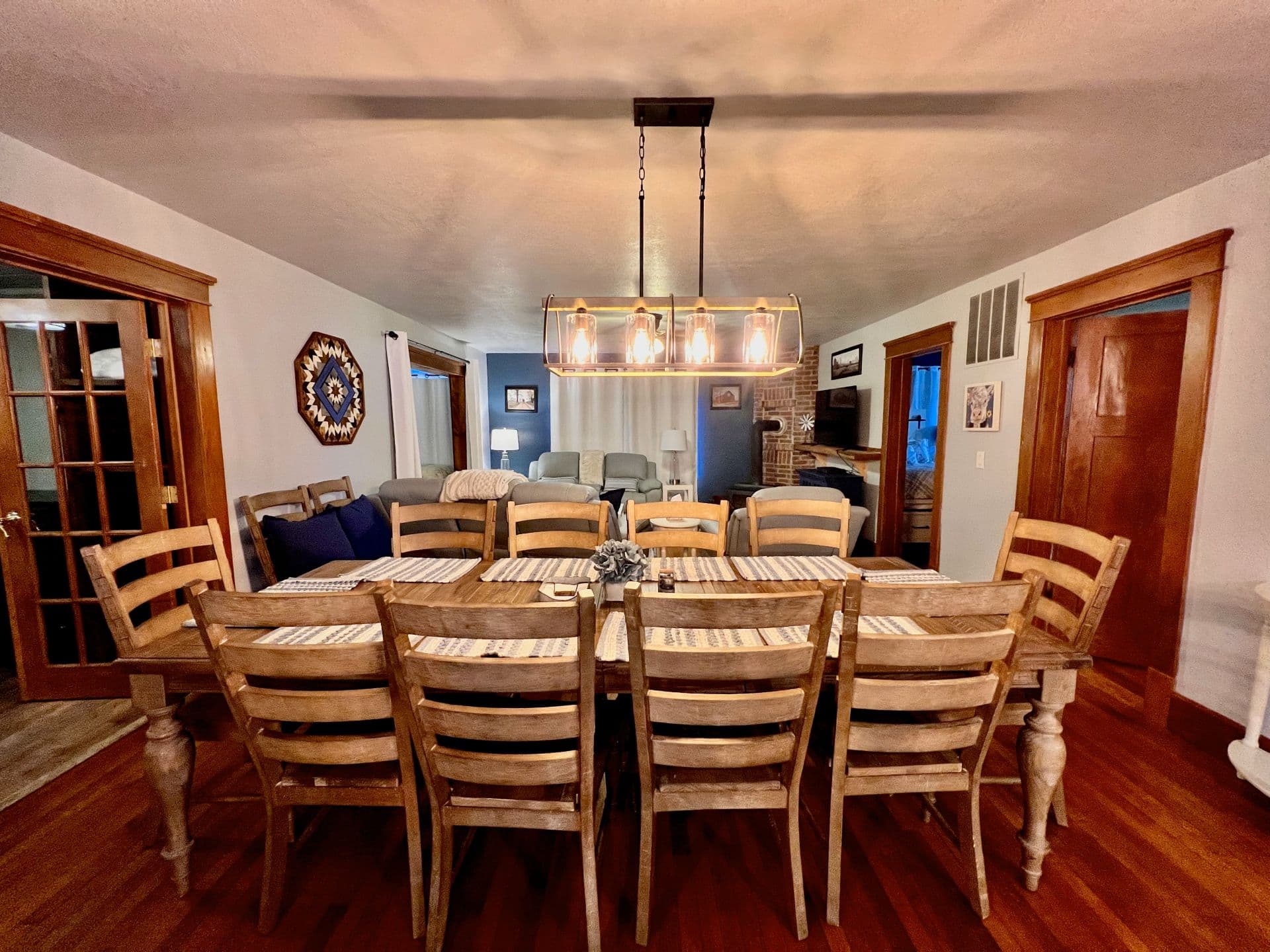 A spacious dining room with a long wooden table and a modern chandelier above, featuring wooden furniture and warm colors.